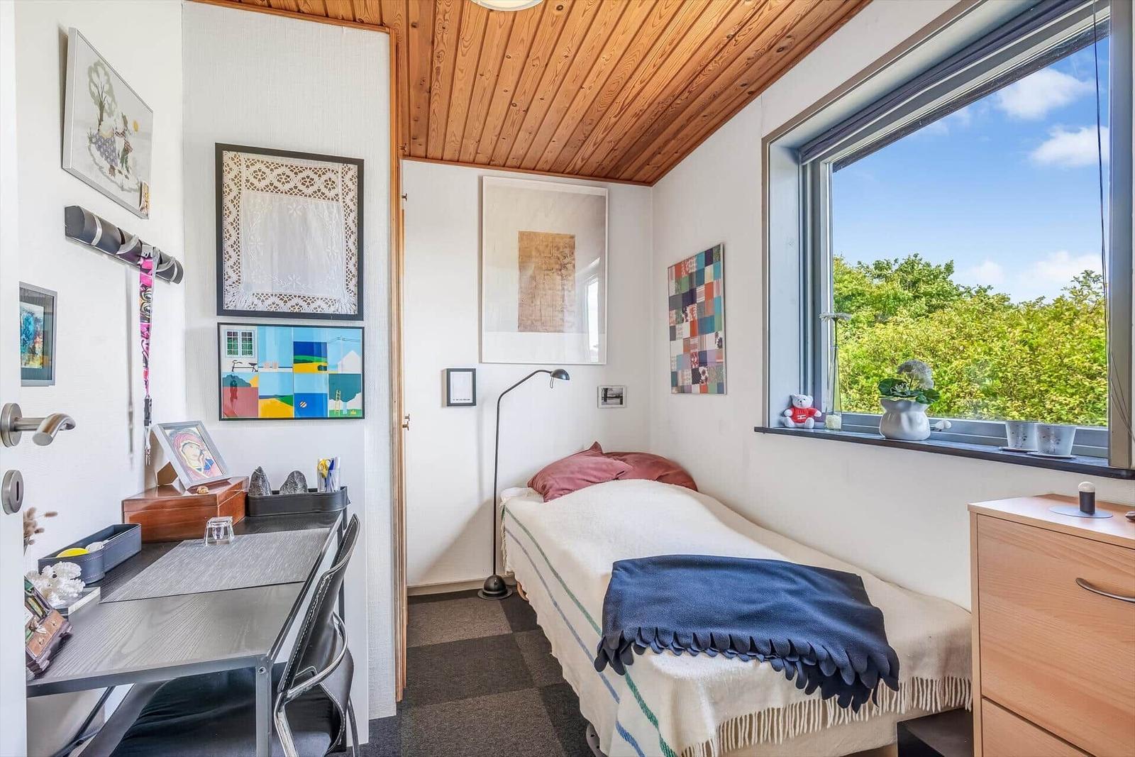 Schlafzimmer mit Bett, Schreibtisch und Fensterblick auf Grün.
