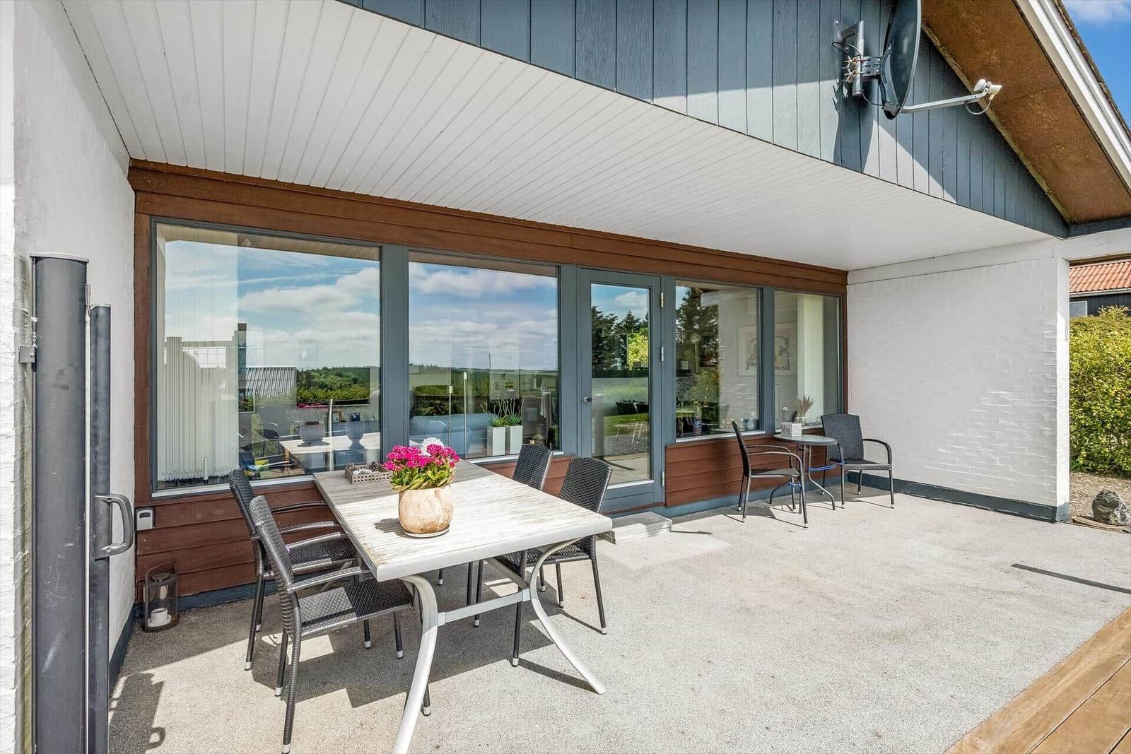 Terrasse mit Tisch, Stühlen und Blumen. Große Fensterfront zum Innenraum.