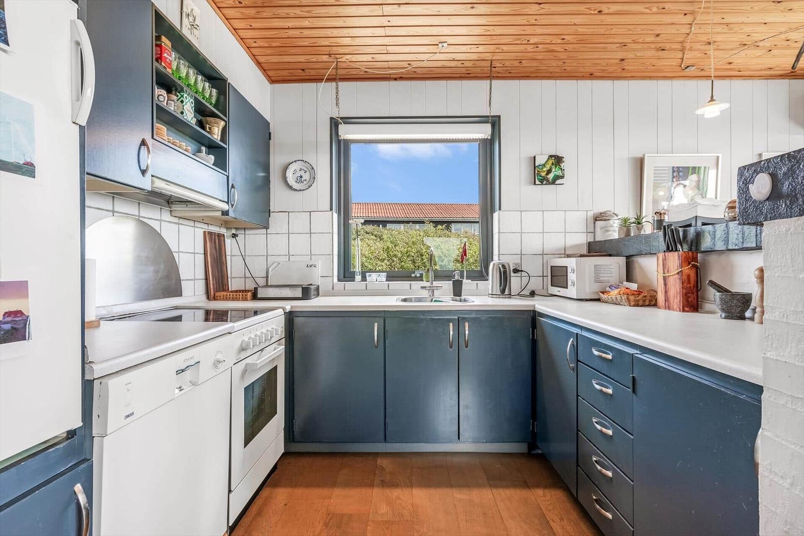 Küche mit blauen Schränken, weißer Arbeitsplatte und Holzboden. Fenster zeigt Blick auf Garten.