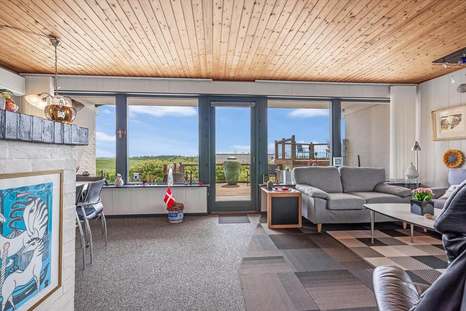 Wohnzimmer mit großem Fensterblick auf Landschaft und Terrasse mit Möbeln.