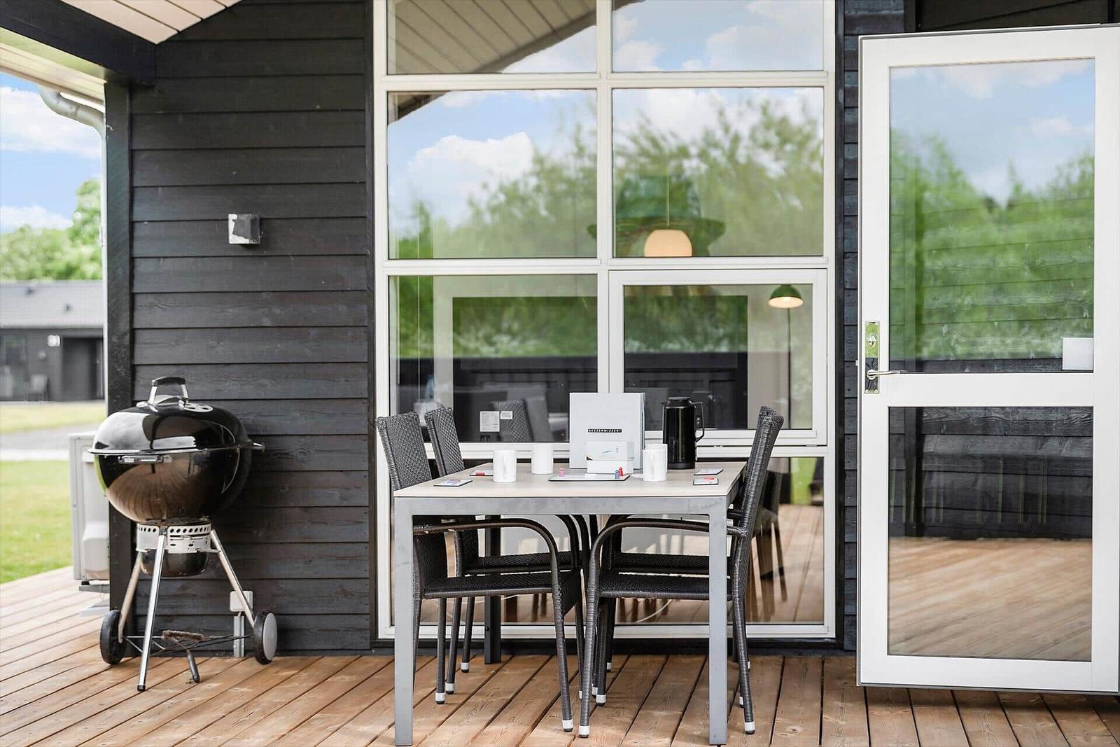 Terrasse mit Grill, Tisch und Stühlen. Große Fenster und Tür führen ins Haus.