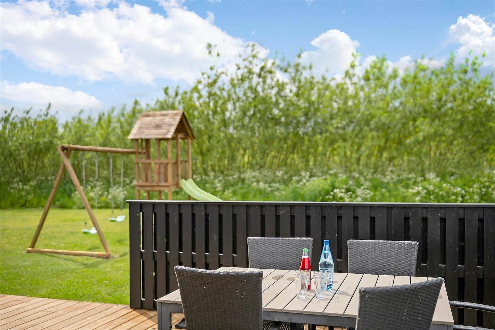 Terrasse mit Tisch und Stühlen, Spielplatz im Hintergrund, grünes Gras und Bäume.