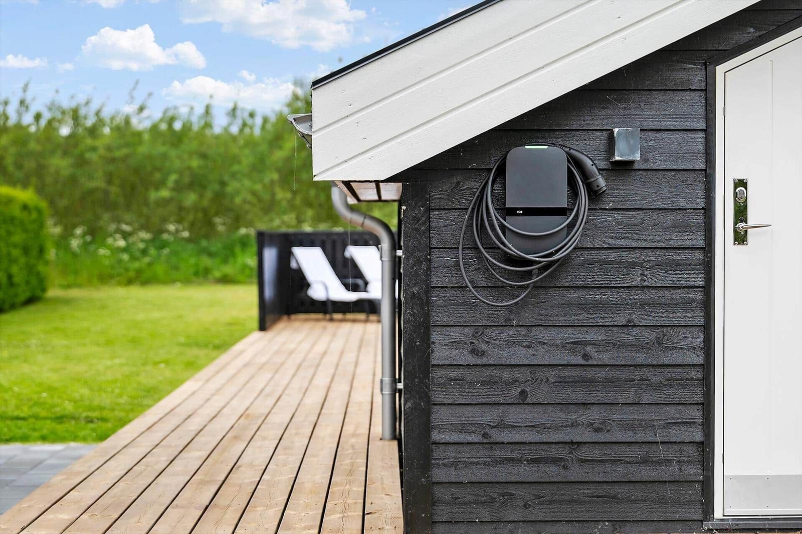Außenansicht eines Holzhauses mit Ladekabel an der Wand und Terrasse mit Liegestühlen.