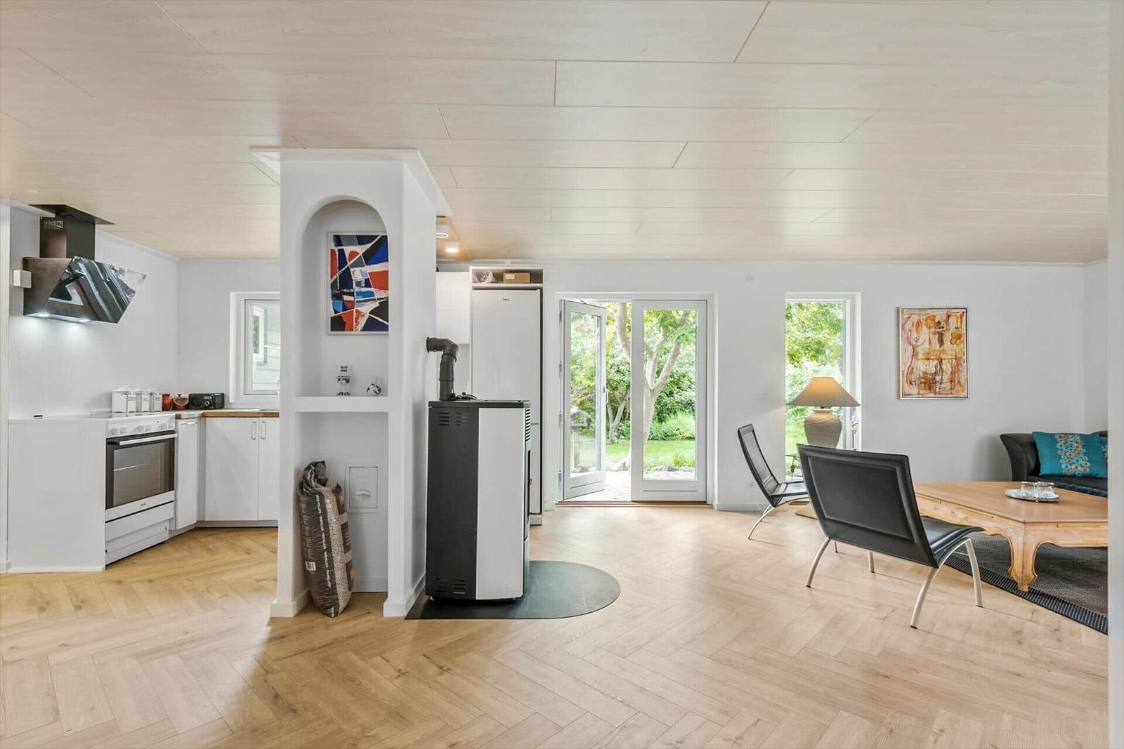 Offener Raum mit Küche, Wohnbereich und Terrassentür. Holzboden und weiße Wände.
