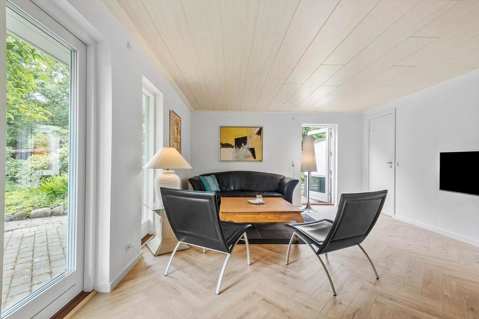 Wohnzimmer mit schwarzer Couch, zwei Stühlen und Holzboden. Große Fenster und Wandbild.