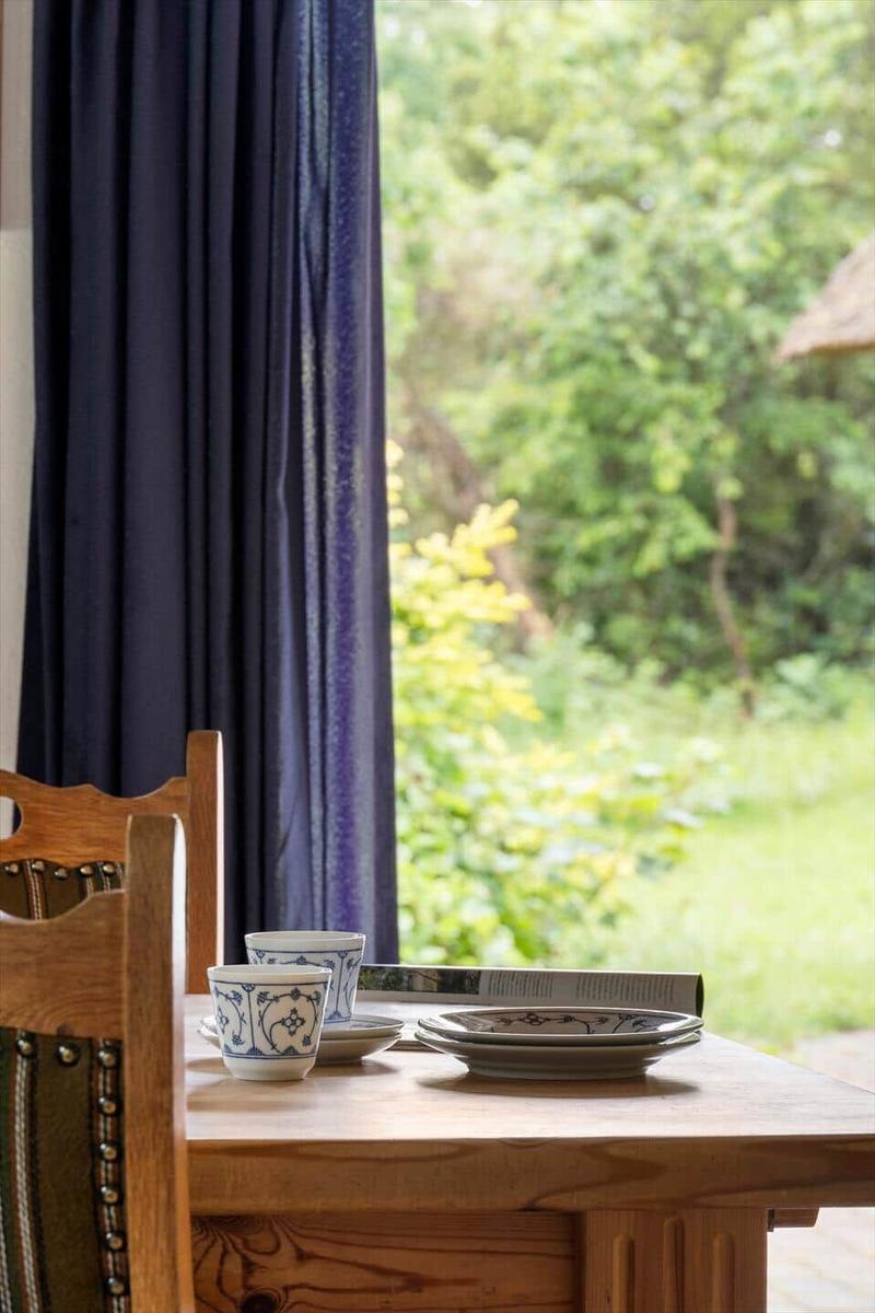 Holztisch mit Tassen und Tellern vor Fenster mit Blick auf Garten.