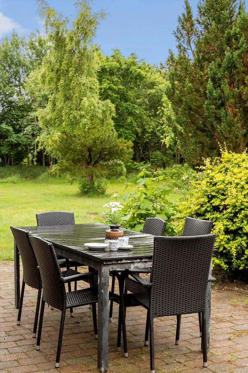 Helle Außenterrasse mit Tisch und Stühlen, umgeben von Grün und Bäumen.