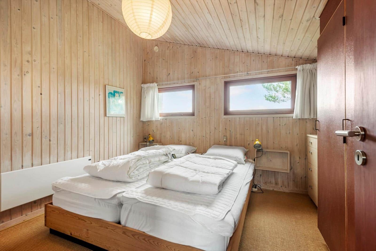 Schlafzimmer mit Doppelbett, Holzwandverkleidung und zwei Fenstern.