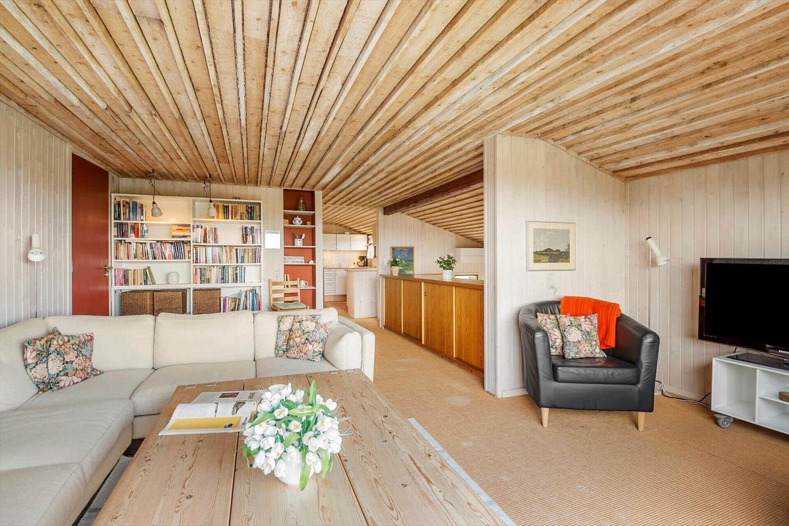 Wohnbereich mit Sofa, Couchtisch und Sessel. Holzdecke und Bücherregale sichtbar.
