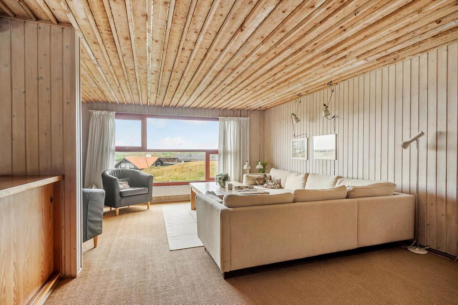 Wohnzimmer mit Holzdecke, Sofa und Sessel. Große Fenster mit Blick auf Landschaft.