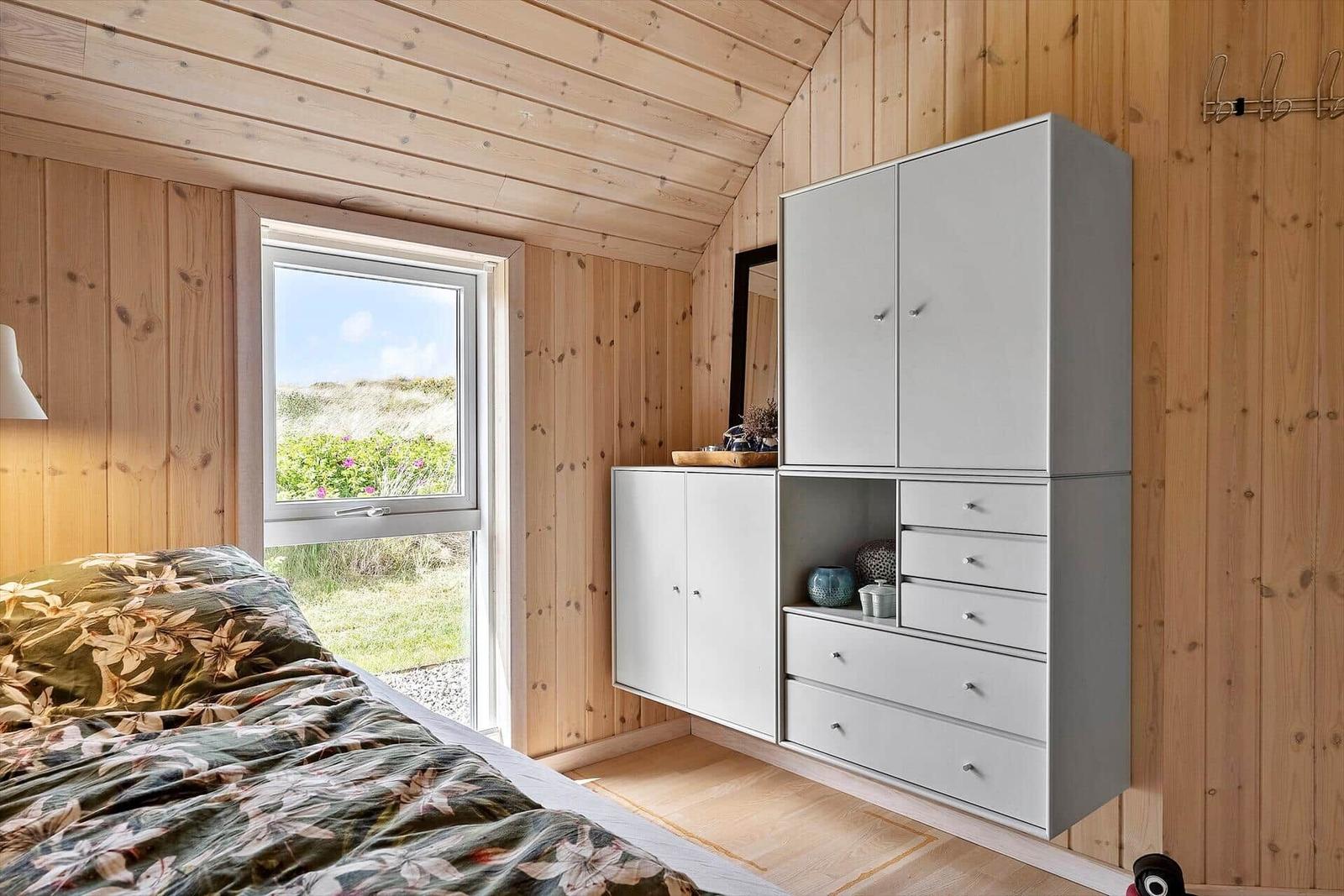 Schlafzimmer mit Holzwand und Fenster zum Garten. Weiße Schrankwand mit Schubladen.