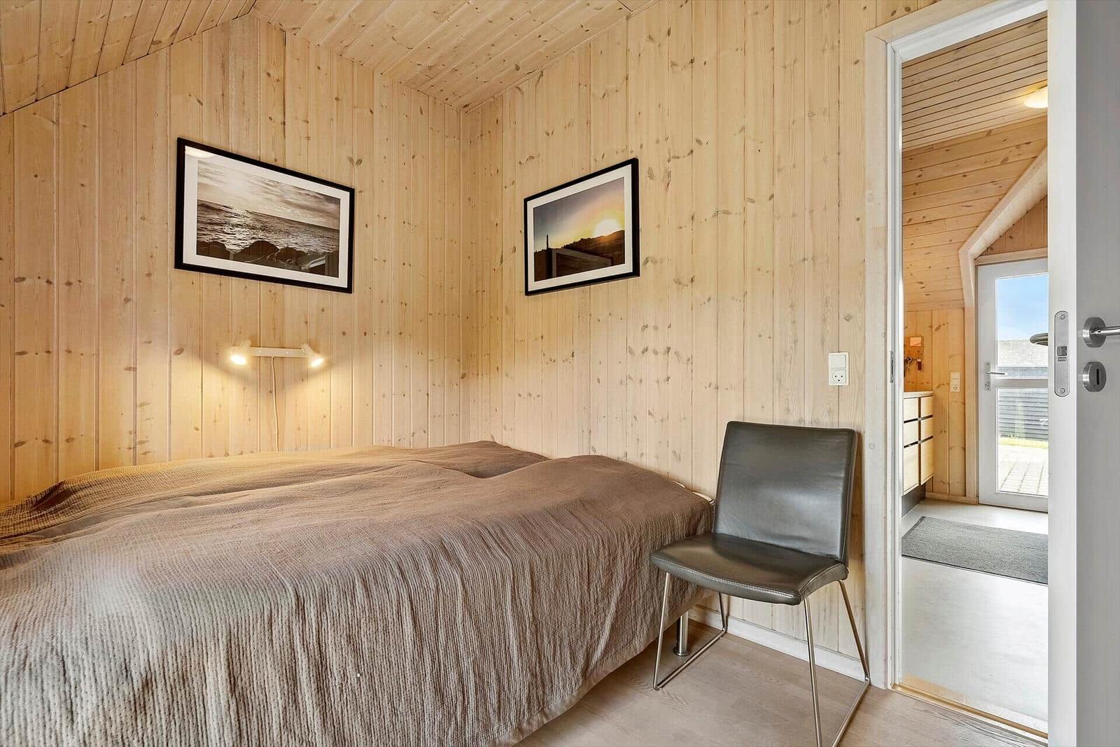 Schlafzimmer mit Holzwand, Bett und Stuhl. Tür führt in Bad.