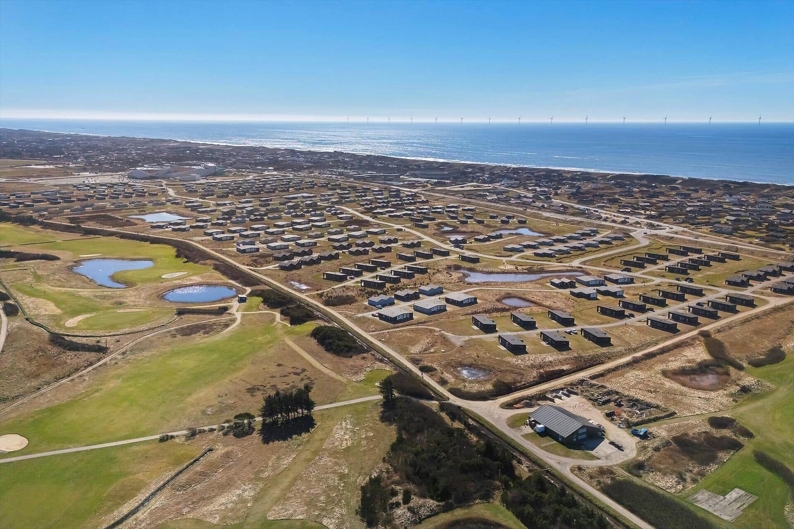 Luftbild von Ferienhäusern am Meer mit Golfplatz und Windkraftanlagen im Hintergrund.