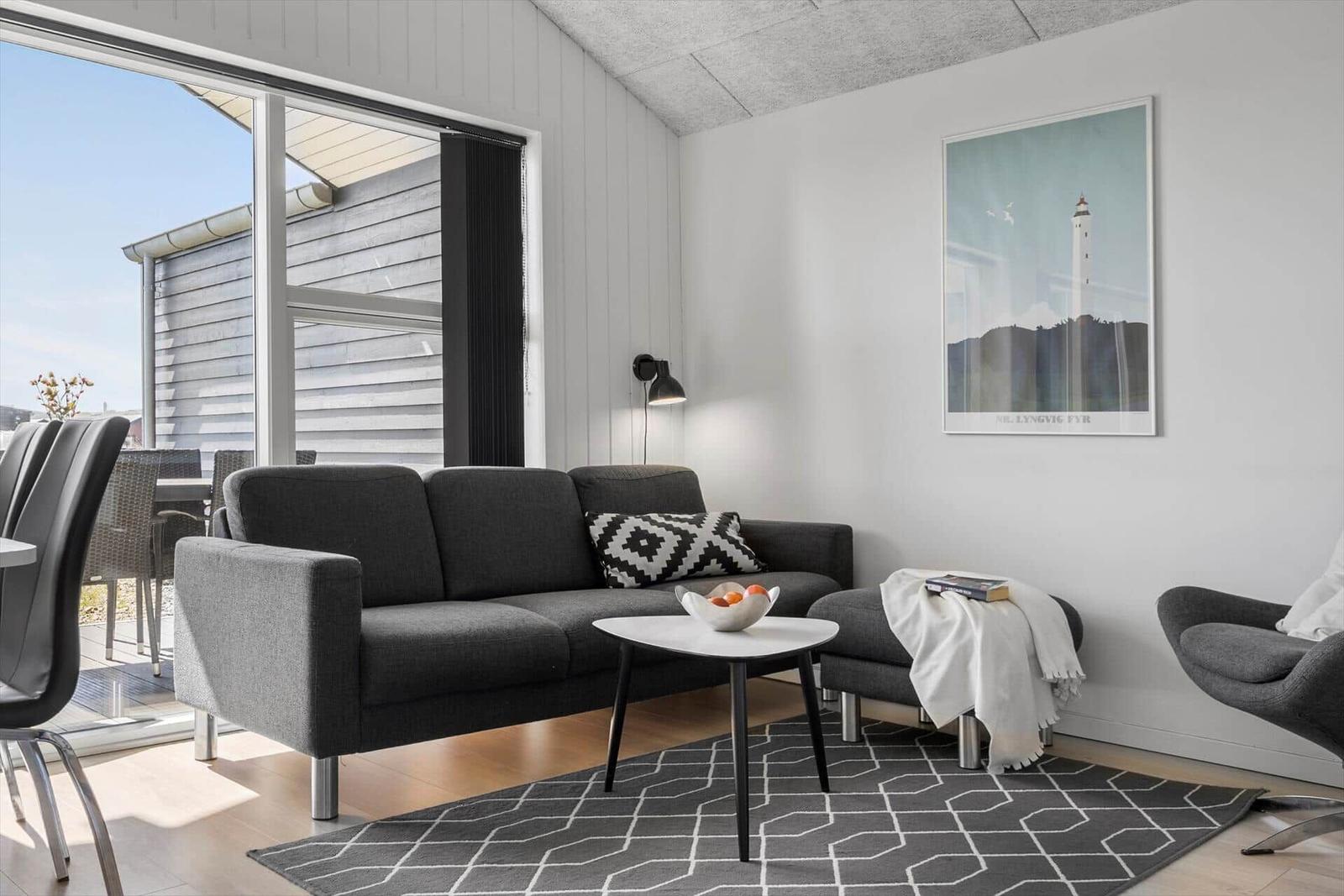 Wohnzimmer mit grauem Sofa, Tisch und Teppich. Große Fensterfront mit Blick auf Terrasse.