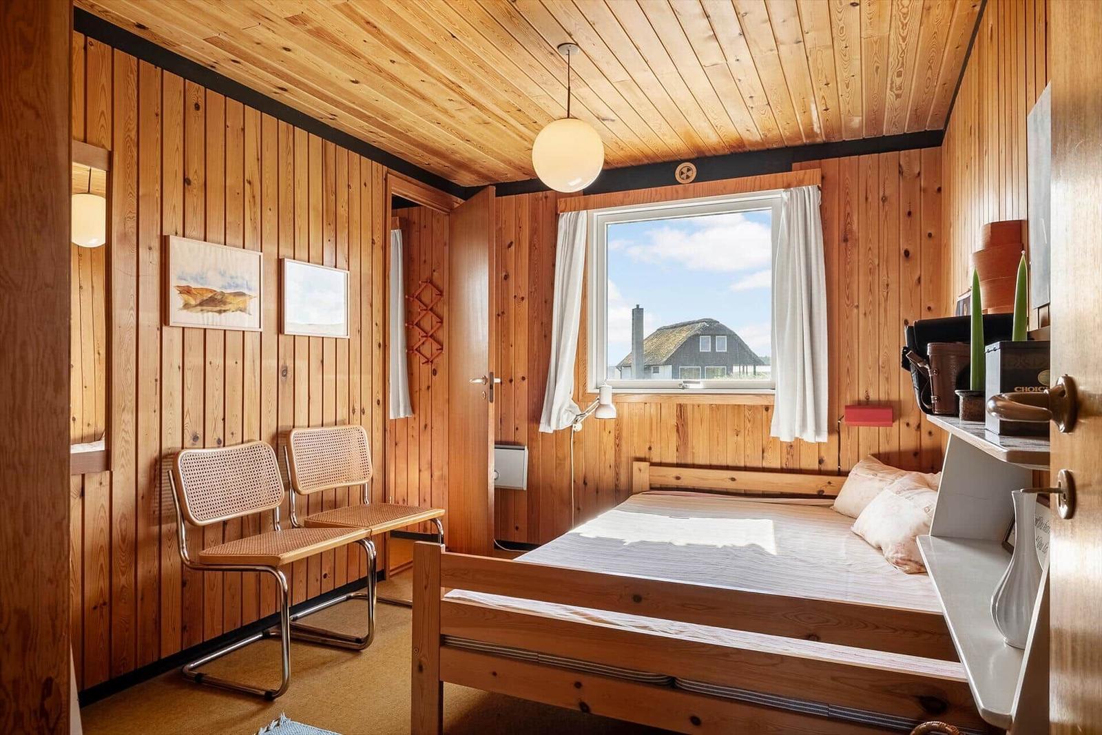 Schlafzimmer mit Holzwand, Bett, Stühlen und Fensterblick auf Haus.