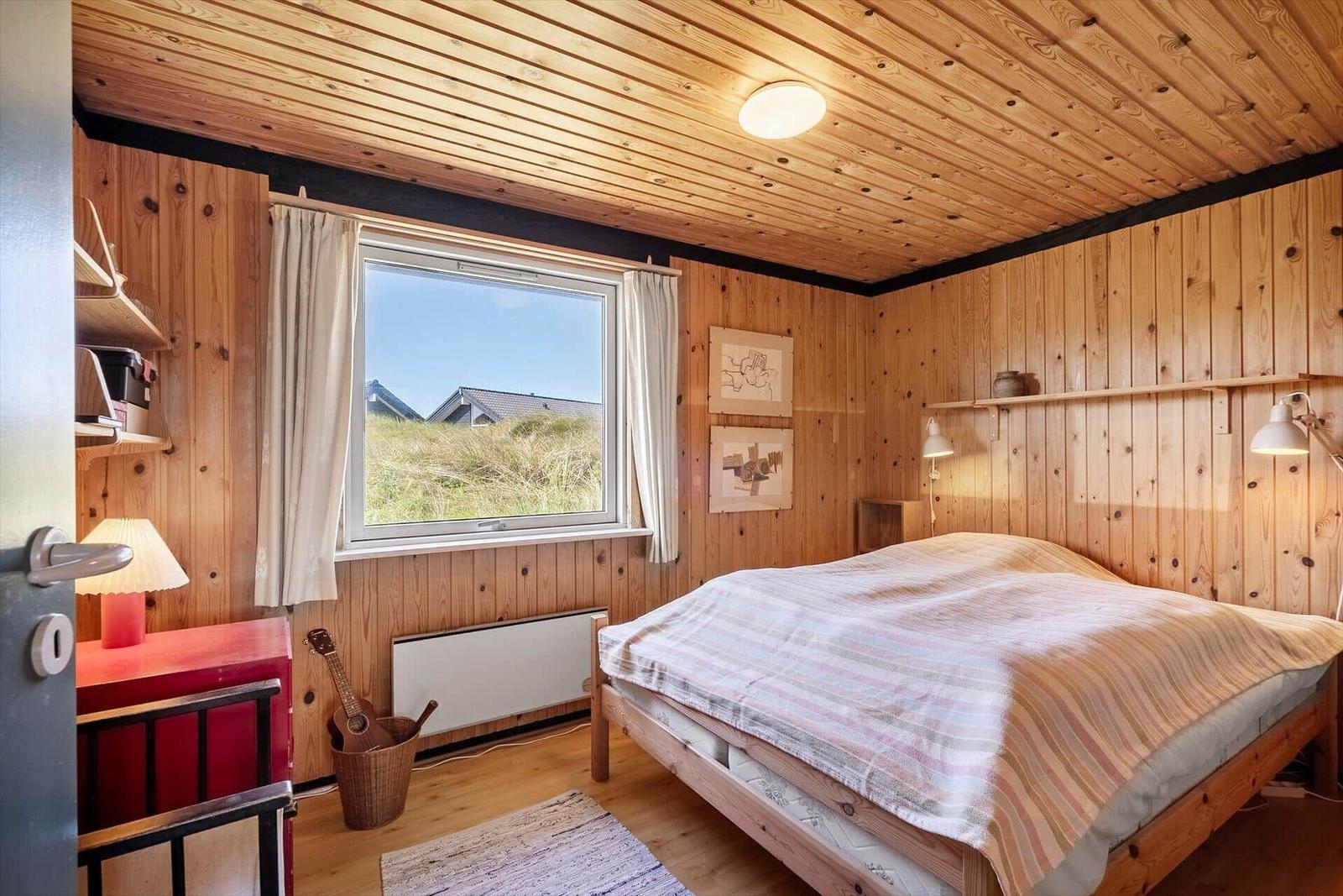 Schlafzimmer mit Holzwand und -decke, Bett, Fenster mit Blick auf Gras und Haus.