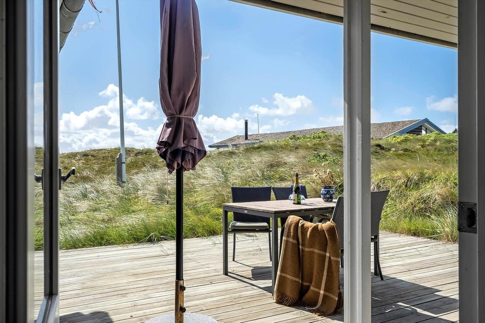 Terrasse mit Tisch, Stühlen und geschlossenem Sonnenschirm. Grasland und Haus im Hintergrund.