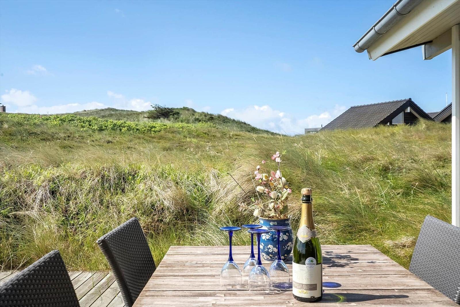 Holztisch mit Flasche und Gläsern auf Terrasse vor Grashügeln.