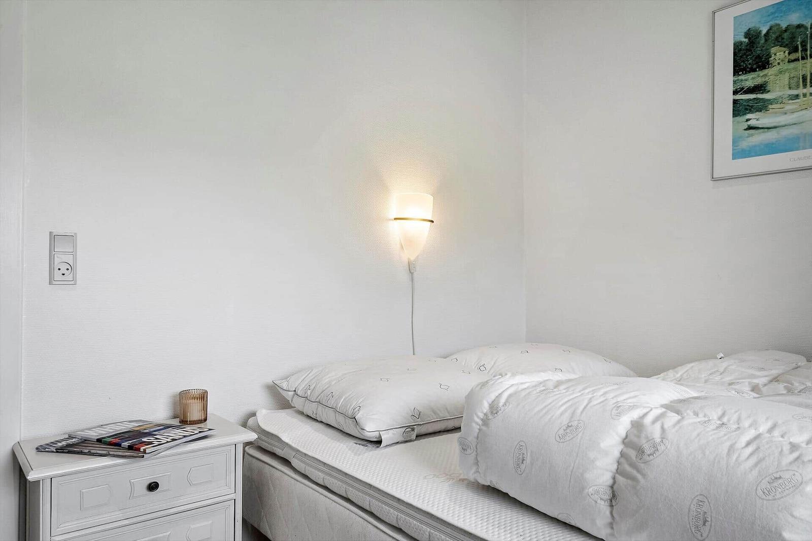 Schlafzimmer mit Bett, Nachttisch und Wandlampe. Auf dem Nachttisch liegen Zeitschriften.