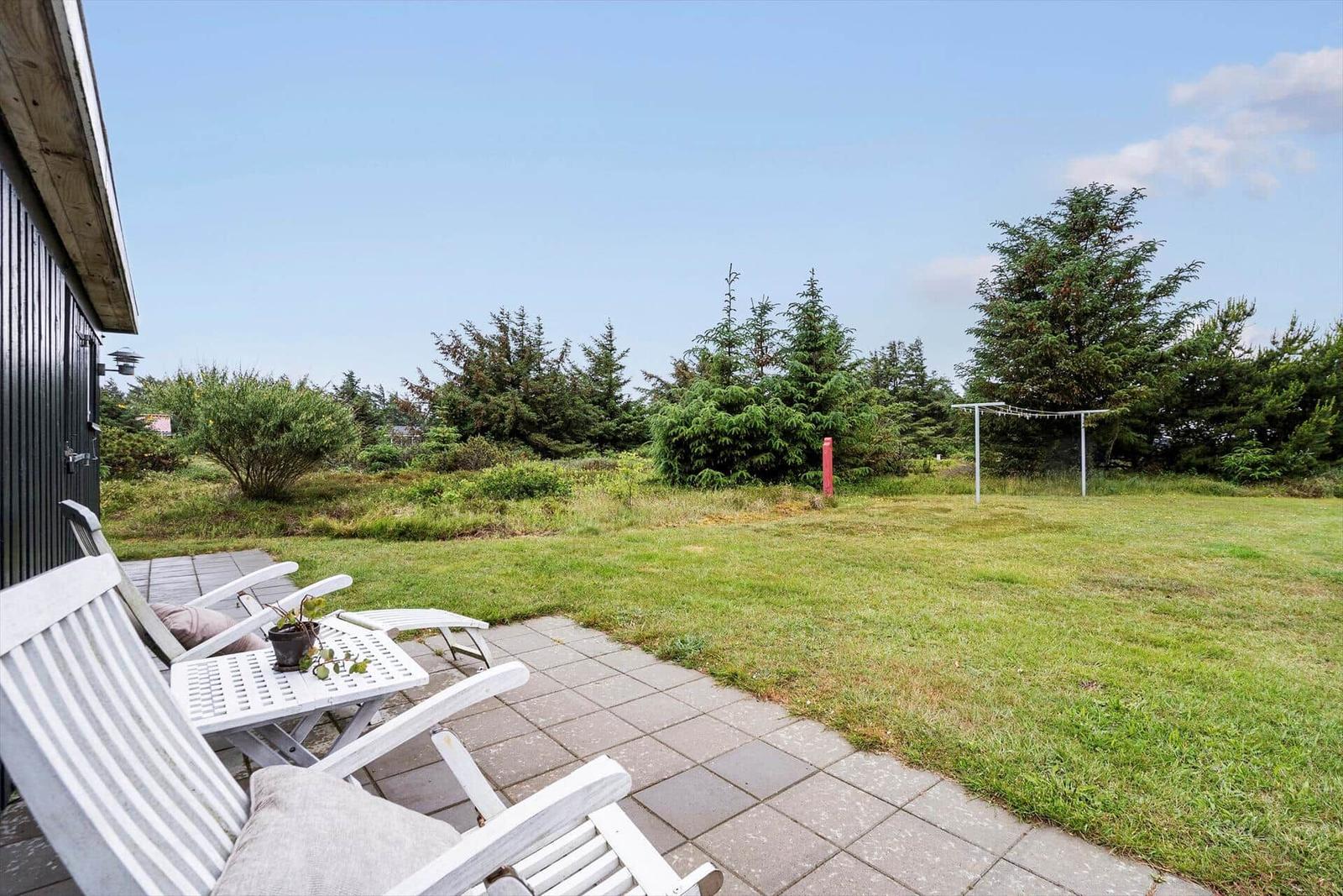 Terrasse mit weißen Stühlen und Tisch, Grünfläche und Bäume im Hintergrund.