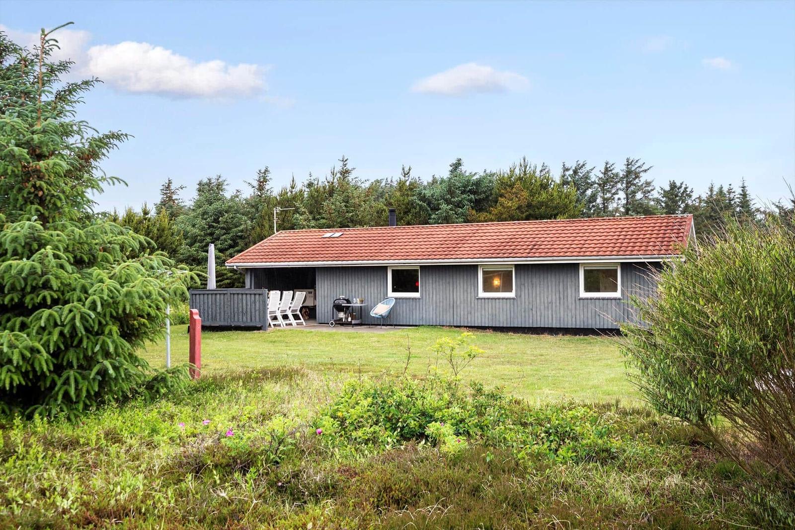 Ein graues Haus mit rotem Dach, umgeben von Gras und Bäumen, mit Terrasse und Gartenmöbeln.