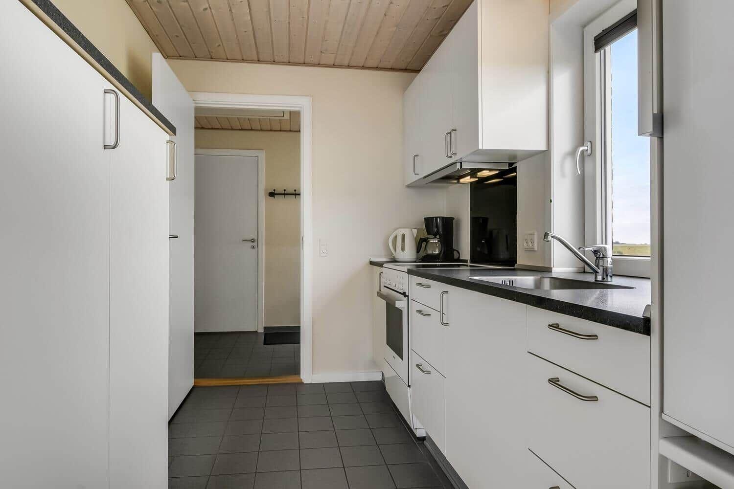 Küche mit weißen Schränken, Arbeitsplatte und Fenster. Durchgang zu einem Raum mit Tür.