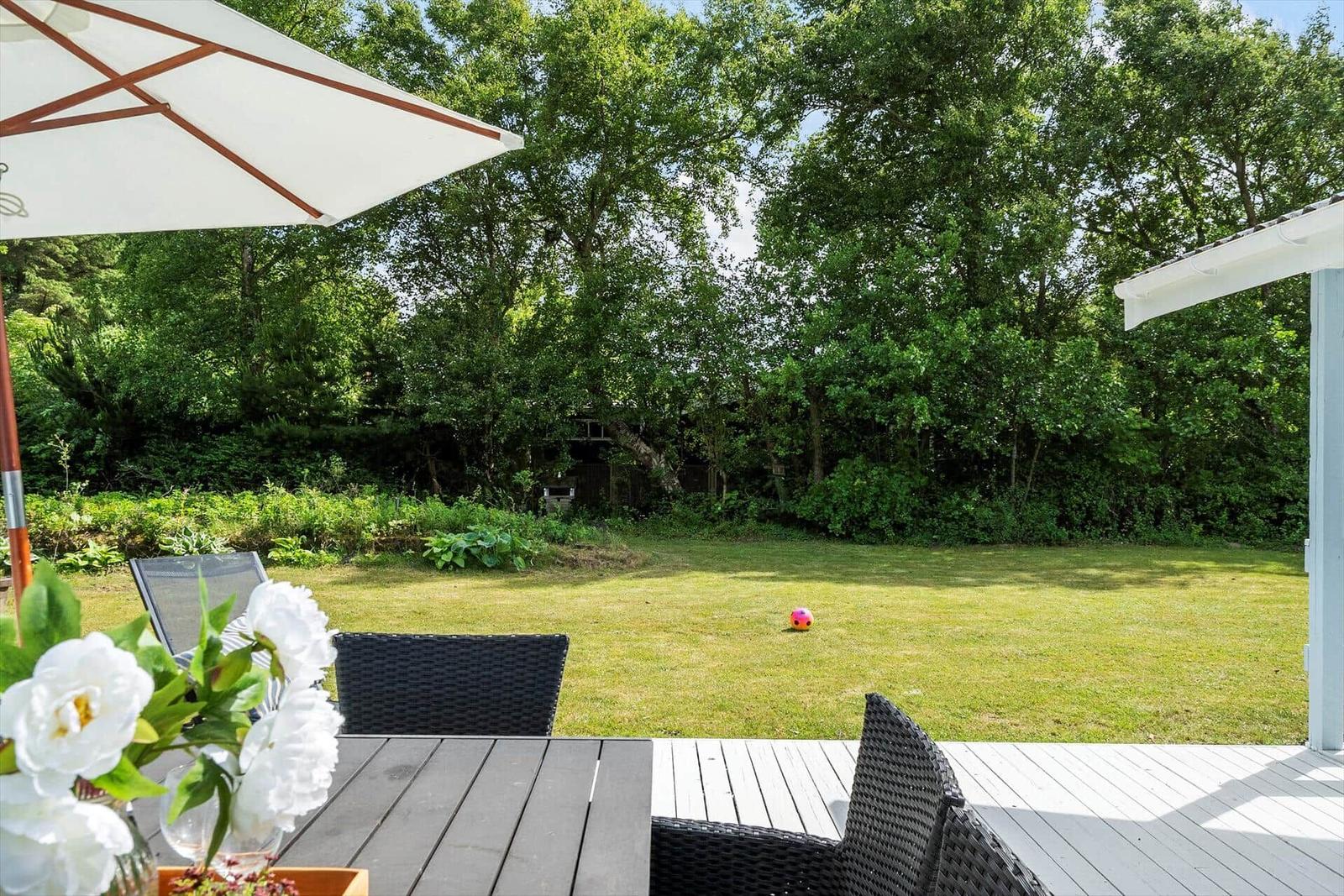 Terrasse mit Tisch, Stühlen und Sonnenschirm. Grasfläche und Bäume im Hintergrund.