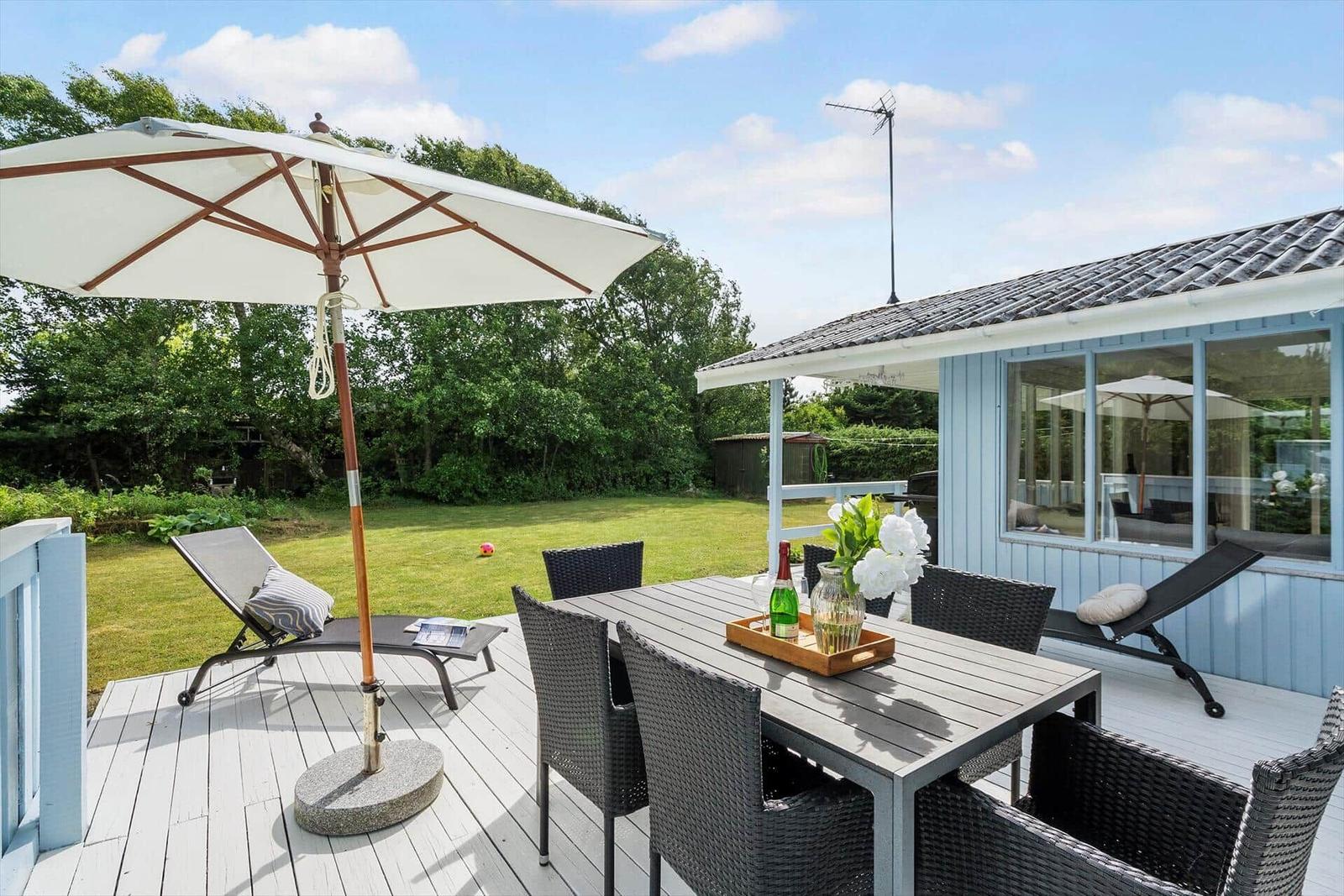 Terrasse mit Tisch, Stühlen und Sonnenschirm. Hintergrund: grüner Garten und Haus.