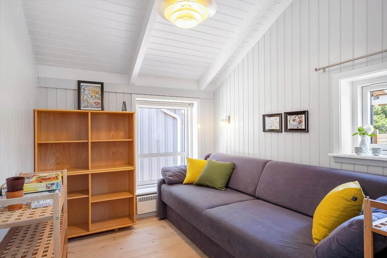 Ein Wohnzimmer mit grafer Couch, gelben und grünen Kissen, Holzregal und weißen Wänden.