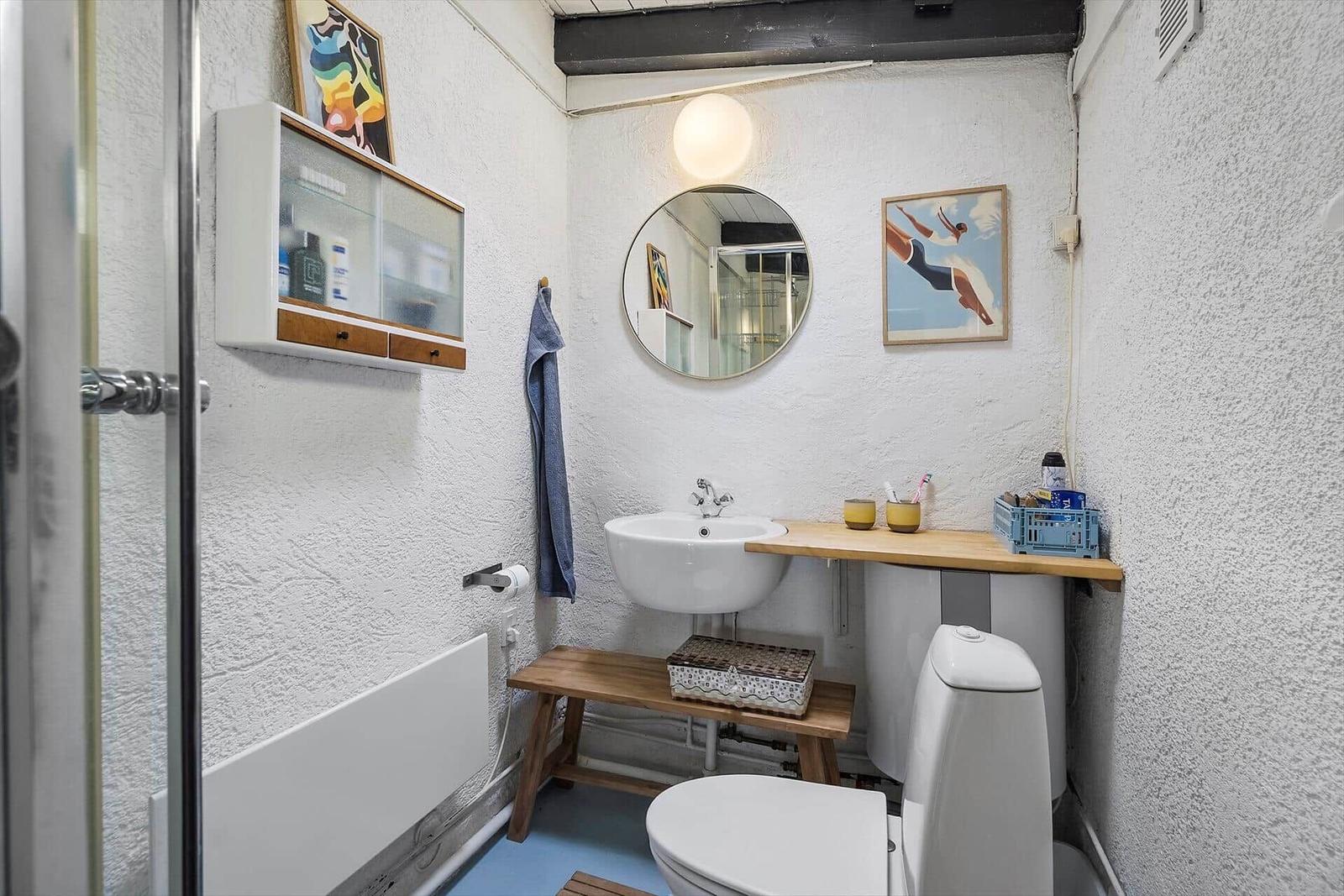 Bathroom with sink, toilet, and shower. Wall art and wooden shelf.