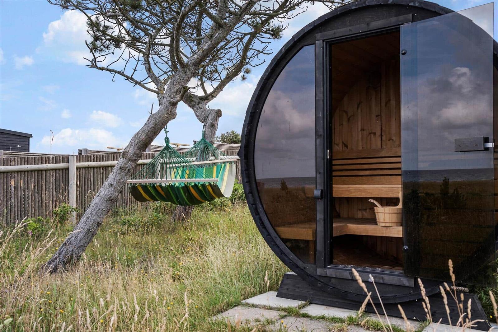 Round sauna with open door and wooden bench. Next to it, a green hammock hangs from a tree.