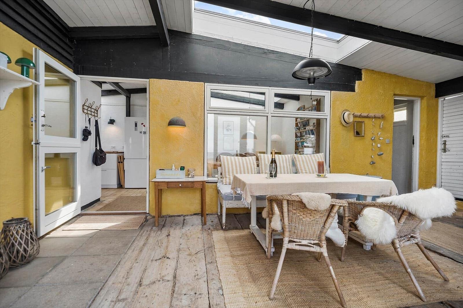 Terrace with dining table, chairs, and view into kitchen. Yellow walls and wooden floor.