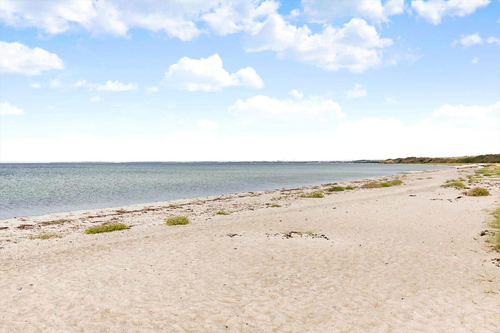 Beach with sand and grass, calm water and blue sky with white clouds.