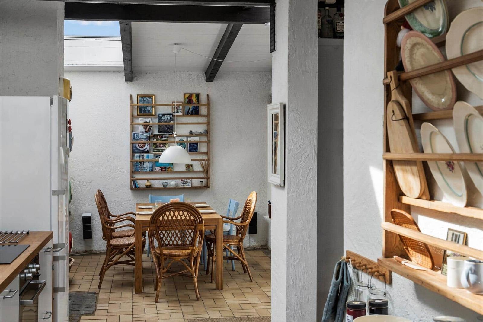 Dining area with wooden table, chairs, and wall shelf. Kitchen with refrigerator and wooden cabinet.