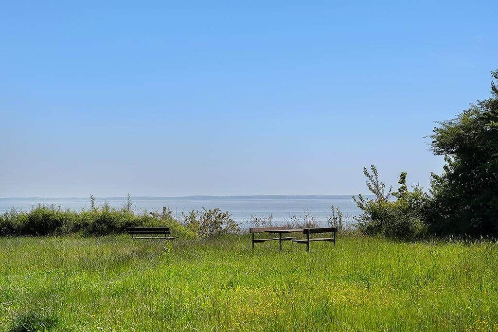 Zwei Bänke stehen in einem Grasfeld am Ufer eines Sees unter blauem Himmel.