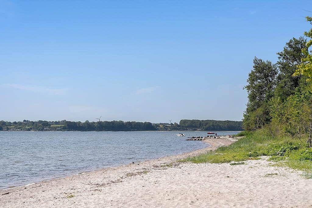 Strand mit Sand und Gras, Wasser im Hintergrund, Bäume rechts, Windräder am Horizont.