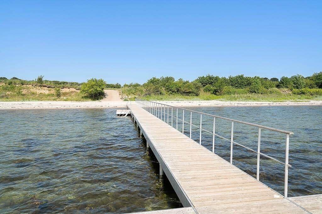 Holzsteg führt über klares Wasser zu einer sandigen Uferstrecke mit Bäumen.