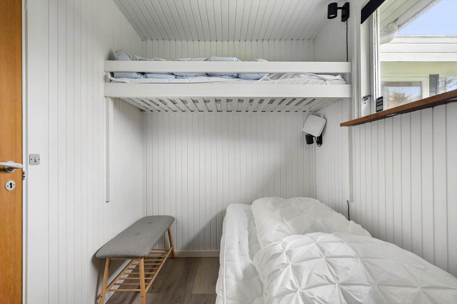 Zimmer mit Doppelbett und oberem Bett, weiße Wände, Fenster und Holztür.