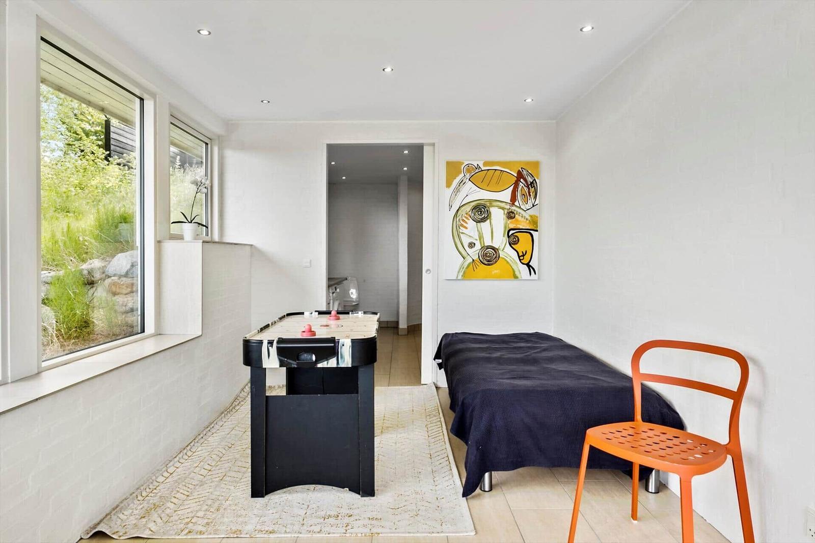 Room with bed, table, and windows. Wall art and orange chair.
