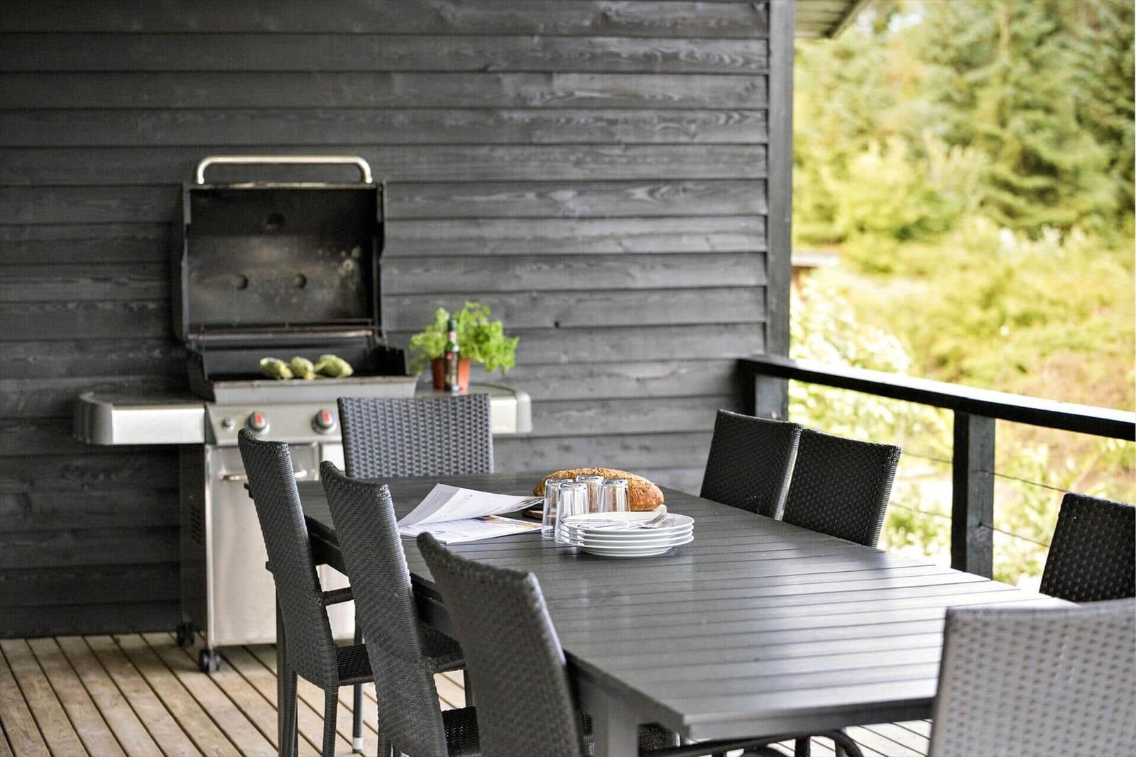 Deck with grill, table, and chairs. Greenery in the background.