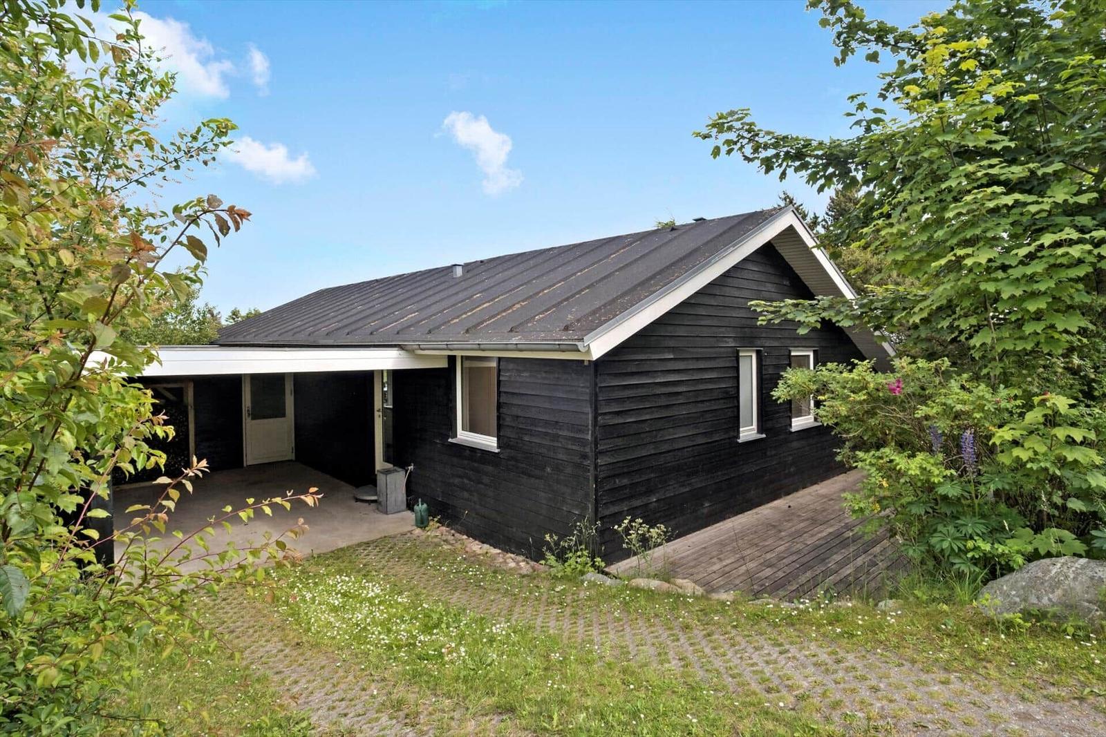 Black wooden house with covered porch and wooden terrace surrounded by greenery.