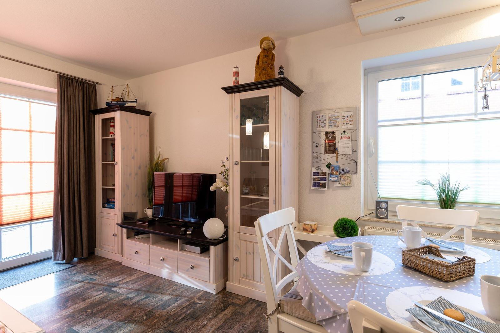 Essbereich mit runden Tisch und weißen Stühlen, daneben Wohnbereich mit TV und Schränken.