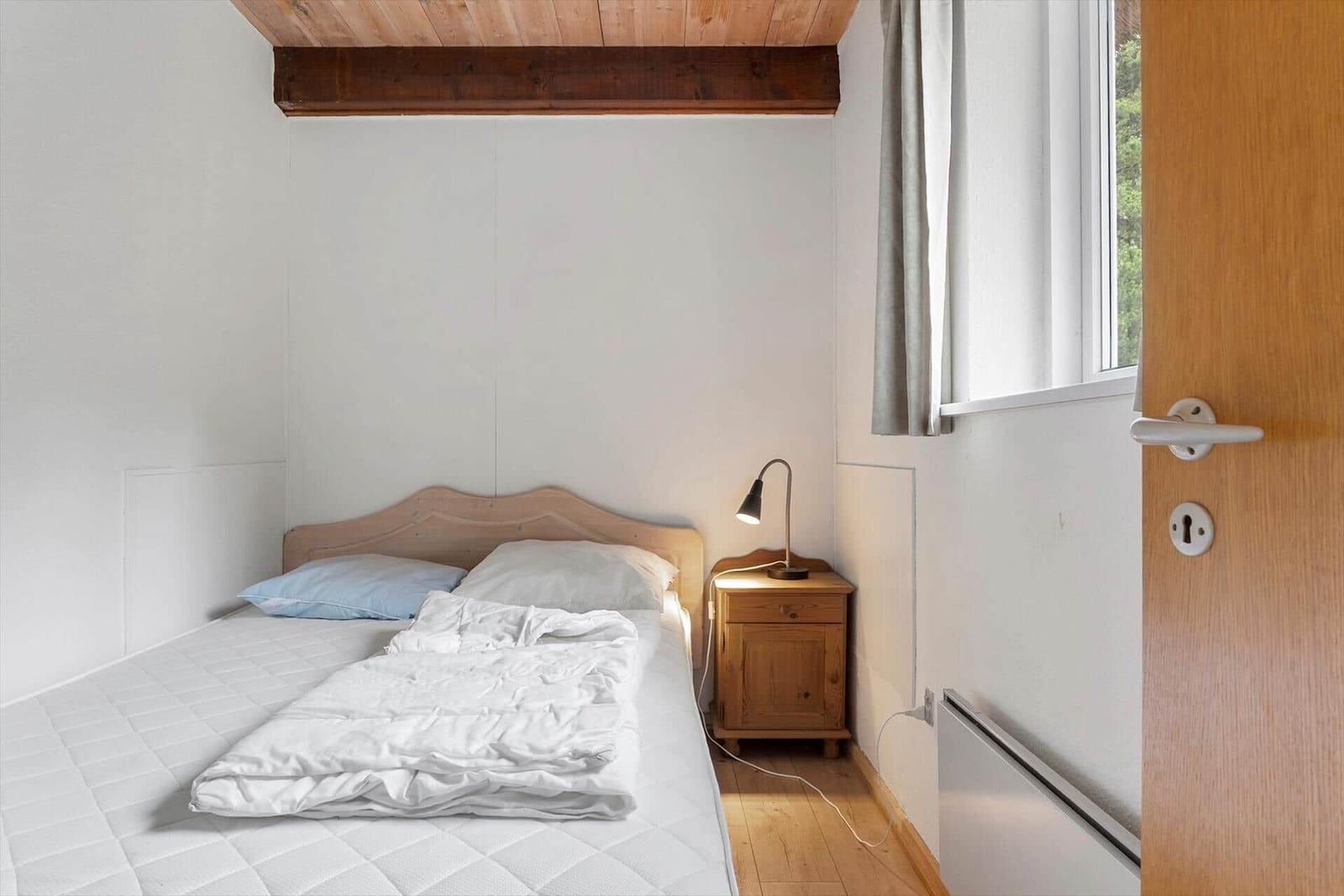 Schlafzimmer mit Bett, Nachttisch und Fenster. Holzdecke und Tür sichtbar.