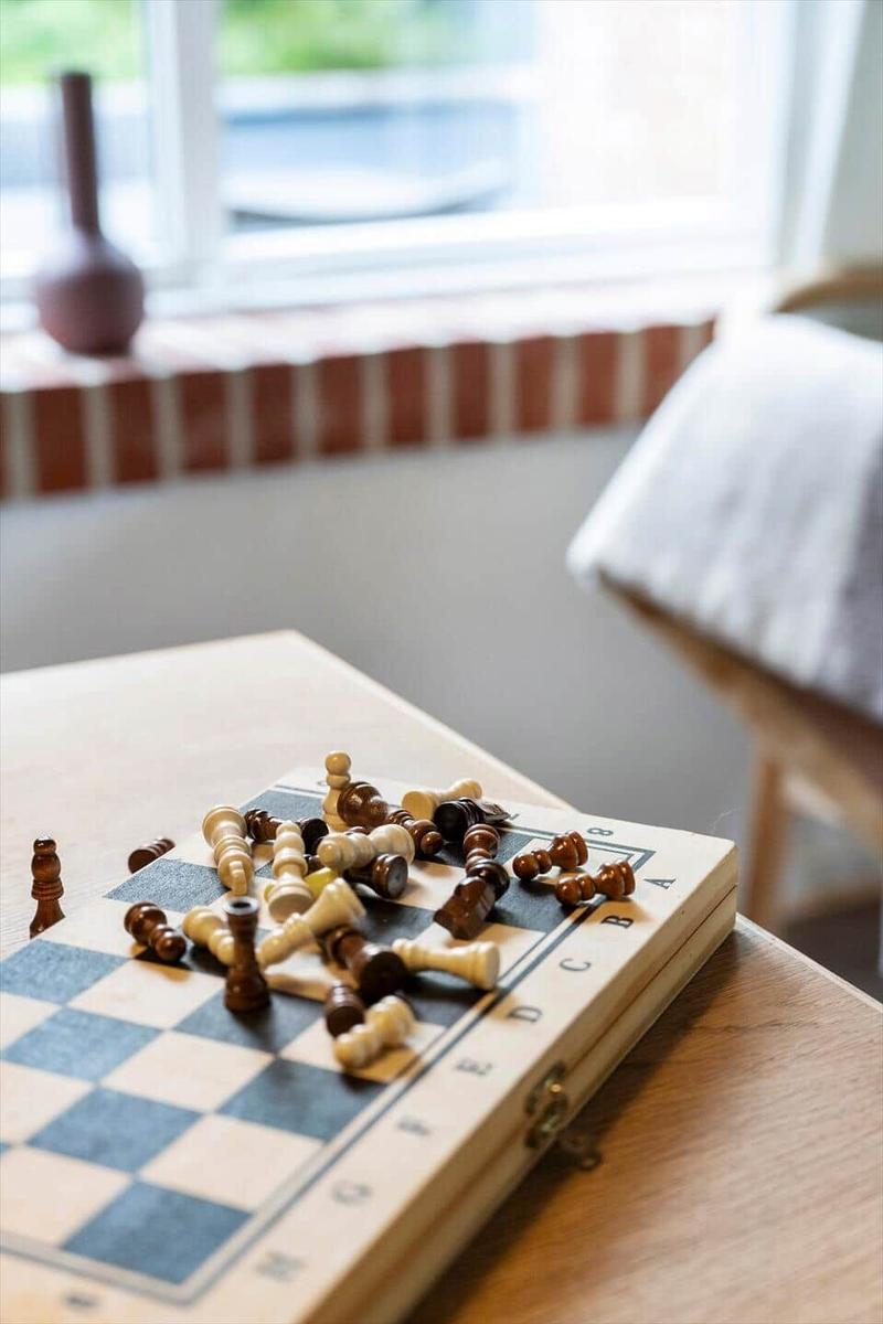 Schachbrett mit Figuren auf Tisch, Fenster und Stuhl im Hintergrund.