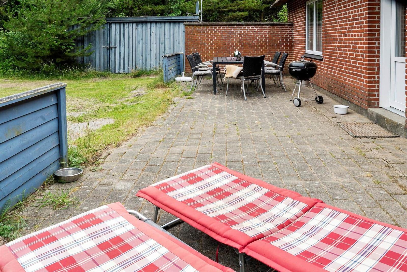 Terrasse mit Liegestühlen, Tisch, Stühlen und Grill. Hinterhof mit Holzzaun und Grünfläche.