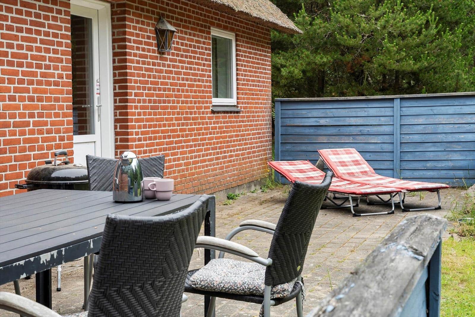 Terrasse mit Tisch, Stühlen, Liegen und Grill neben rotem Backsteinhaus.