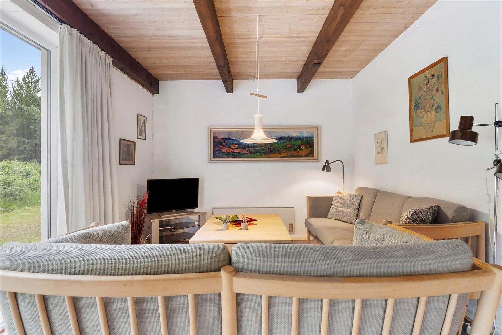 Wohnzimmer mit Holzdecke, Sofa und Fernseher. Fenster mit Blick auf Wald.