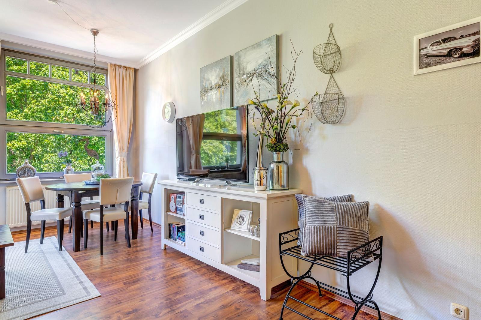 Ein Essbereich mit Tisch, Stühlen und großem Fenster. Fernseher auf weißem Schrank mit Dekoration.