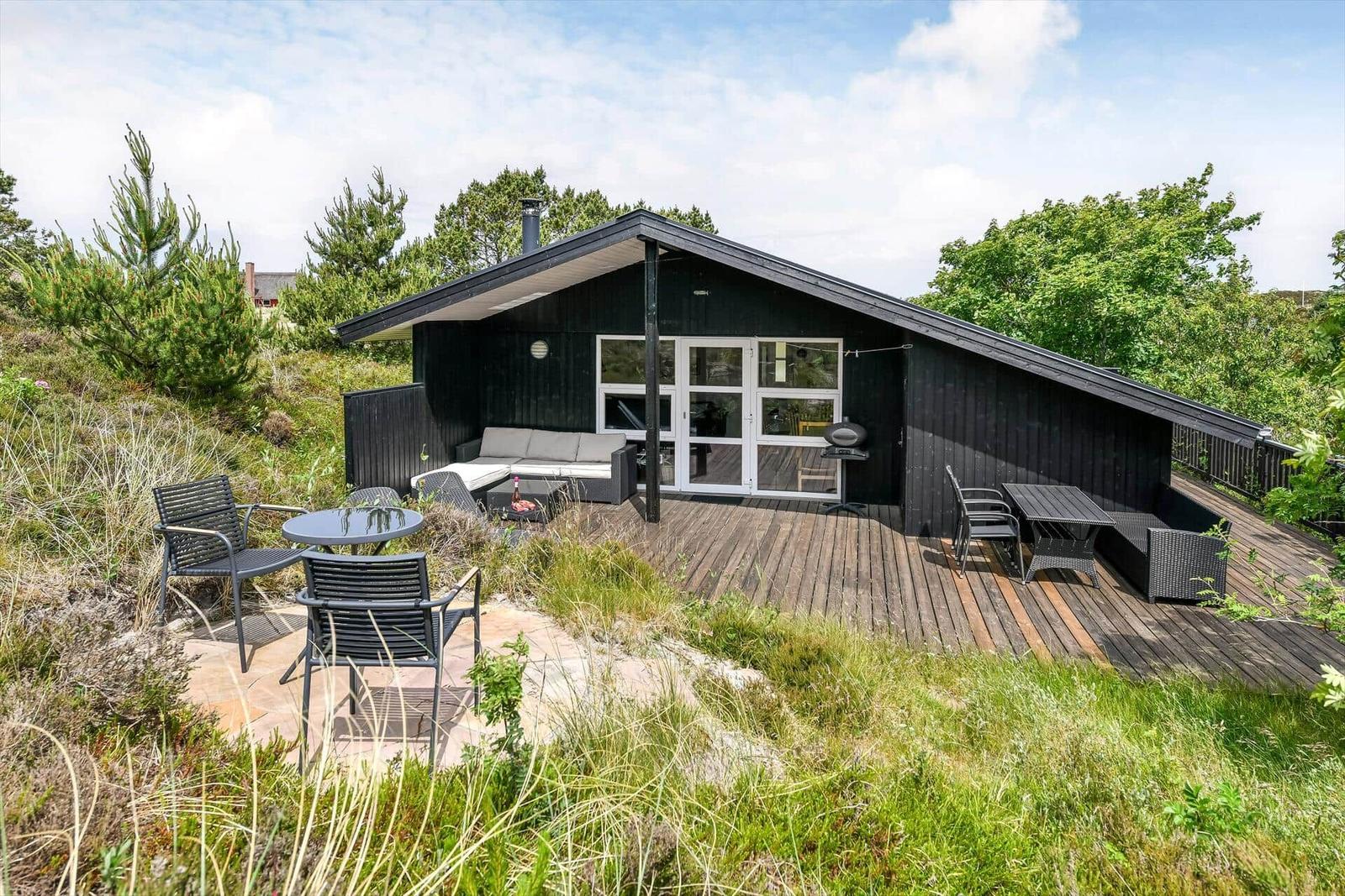 Schwarzes Haus mit Terrasse, Gartenmöbeln und Grill. Umgeben von Gras und Bäumen.