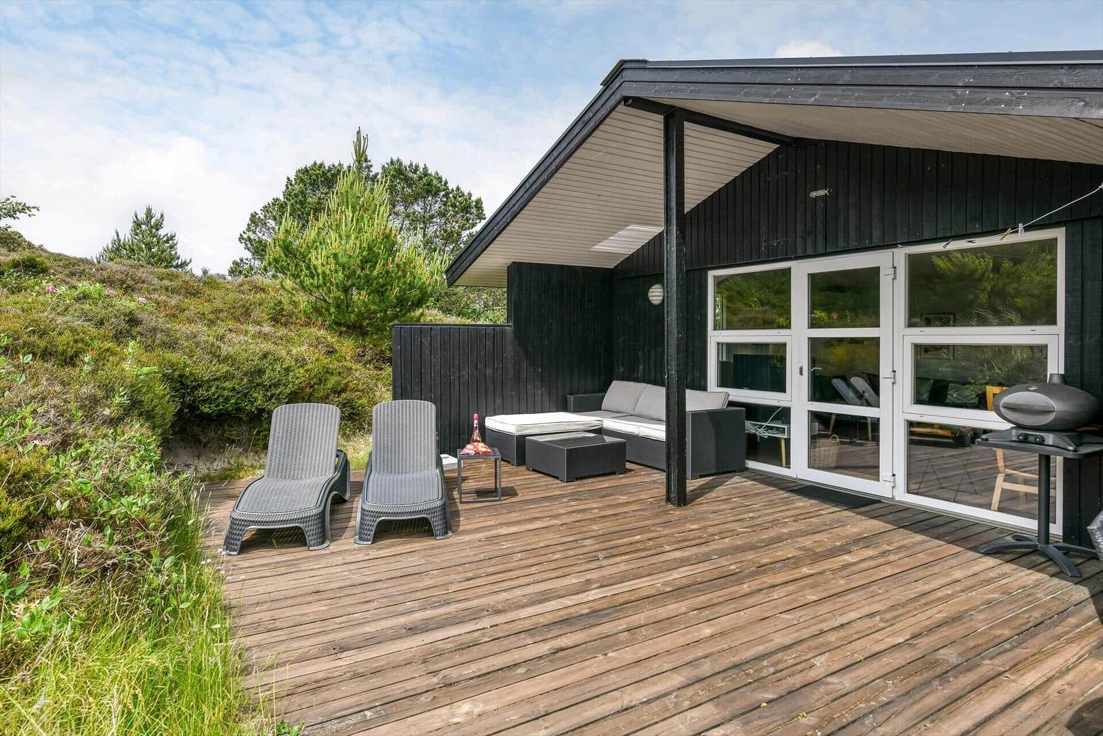 Terrasse mit Liegen, Sofa und Grill. Schwarzes Haus mit Holzdecke und Glasfront.